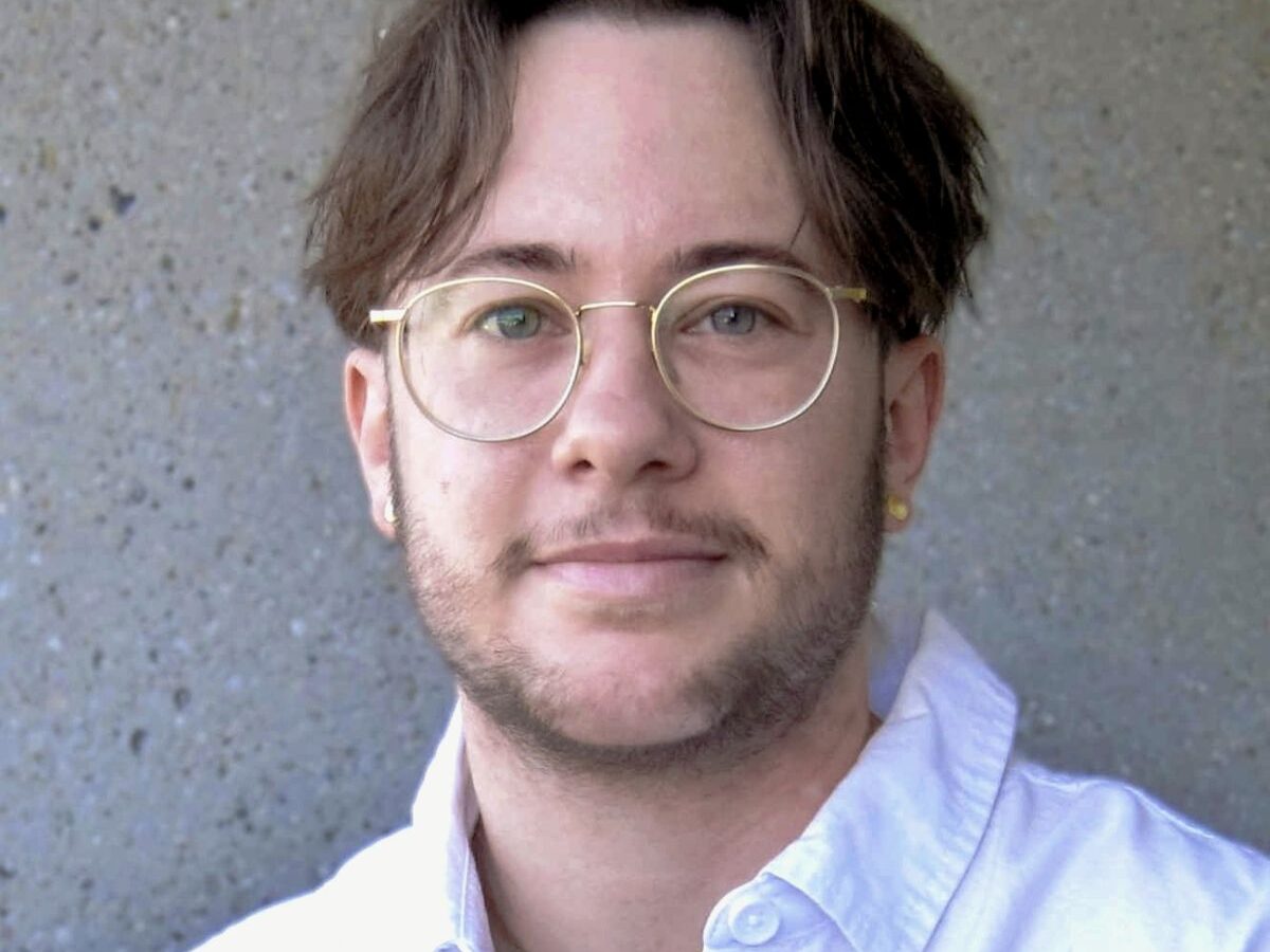 Portrait von Monet Wohlgemuth, er ist eine weisse Person mit braunen Haaren und einem leichten Bart. Er schaut direkt in die Kamera und trägt ein weisses Hemd und eine Brille.